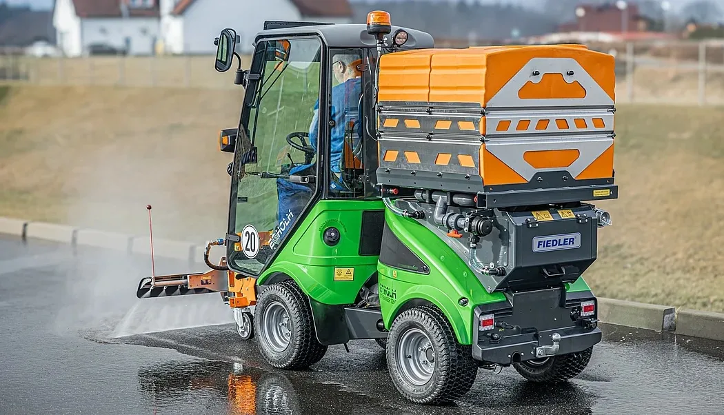 Kompaktes grün-orangefarbenes Fiedler-Fahrzeug wässert eine nasse Wegfläche mit einer Frontsprühleiste