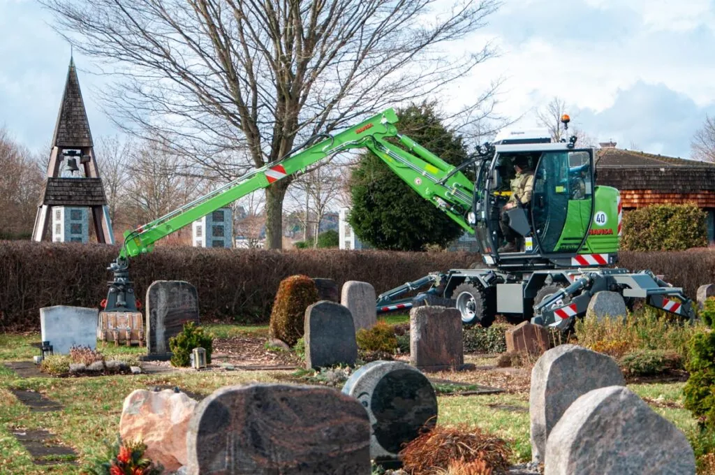 Grüner Hansa Kommunalbagger mit Greifarm auf einem Friedhof